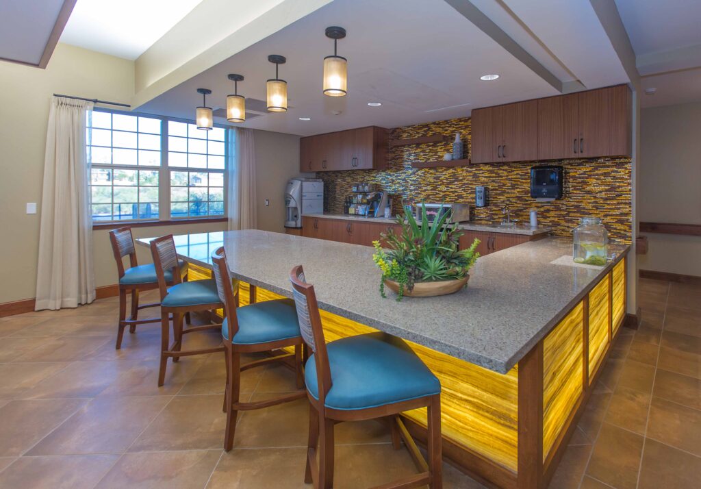 Sage Desert Tucson AZ 2018 June -0331 - The Ranch Estates of Tucson The Ranch Estates of Tucson bar area with a bar table, blue chairs, a potted plant, brown cupboards, and kitchen appliances against the wall.